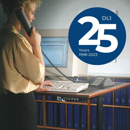 办公室人员在使用 LINAK DL1 系统驱动的电动升降办公桌。DL1 25 年纪念标志：1998 年 - 2023 年