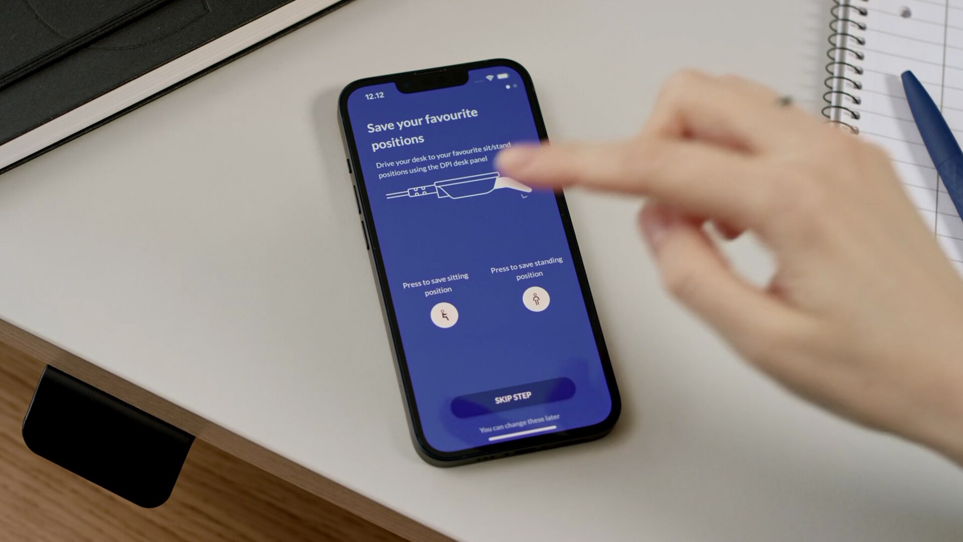 办公桌上的移动设备显示如何通过 Desk Connect™ App 在 LINAK 桌面板 DPI1C 上设置喜爱的坐站姿势屏幕。